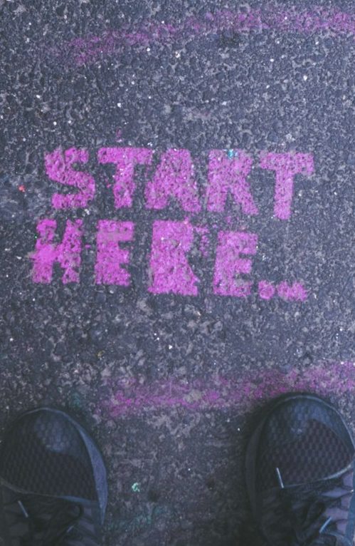 Jetzt in Umsetzung kommen Pink "START HERE" Schriftzug auf dem Boden, sichtbar vor einem Paar Schuhe.